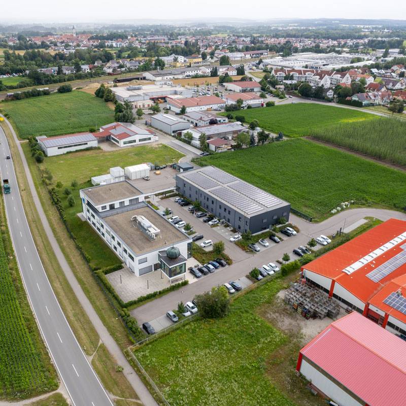 RENG-Industriesysteme-GmbH-126 Luftaufnahme eines modernen Gewerbeareals mit mehreren Büro- und Produktionsgebäuden, großzügigen Parkflächen und umliegenden Feldern. Das Hauptgebäude besitzt einen markanten Eingangsbereich mit verglastem Turm. Die Umgebung zeigt weitere Gewerbehallen, Straßenanbindung und angrenzende Wohngebiete.