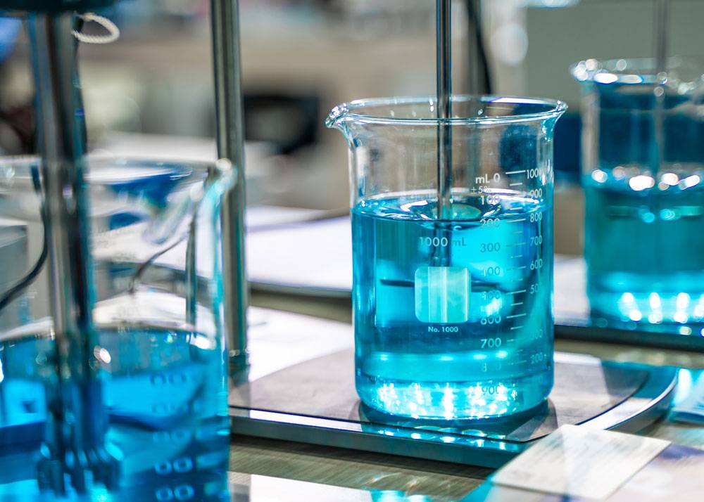 Reng-Ventiltechnik-Chemie-Anwendung Das Bild zeigt ein großes Becherglas mit blauer Flüssigkeit, das auf einer Laborwaage oder einem Rührgerät steht. In der Flüssigkeit befindet sich ein Rührstab, der von oben in das Gefäß führt. Die Skala auf dem Glas ist gut sichtbar und zeigt ein Volumen von bis zu 1000 Millilitern. Im Hintergrund stehen weitere Bechergläser mit ähnlicher Flüssigkeit. Die Szene vermittelt einen typischen Arbeitsschritt in einem chemischen oder analytischen Labor.