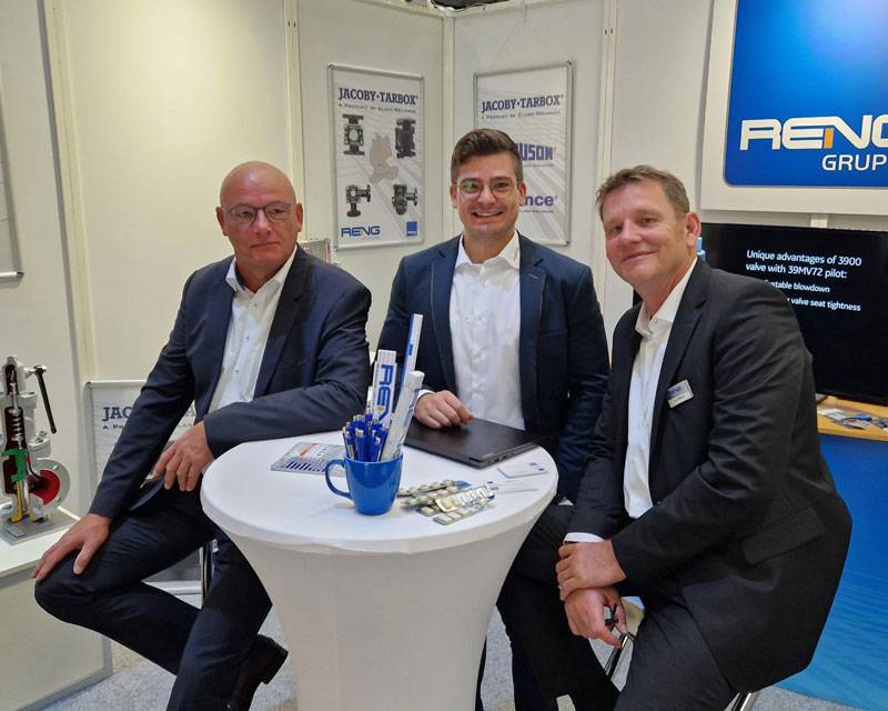 Reng-ventiltechnik-Schulungen Drei Männer in schwarzen Anzügen sitzen auf Barhockern hinter einem Stehtisch auf einem Messestand der Reng Gruppe. Der linke Mann blickt ernst in die Kamera, während der mittere Mann in die Kamera grinst und der rechte nur schmunzelt.